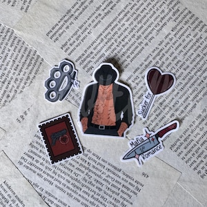 Op de afbeelding: Een set van vijf stickers met een boksbeugel, een persoon met een hoed en open jas, een hartvormige lolly met de tekst Sucker for, een rechthoekige postzegel met een pistool en hart, en een mes met de tekst Mafia Romance.