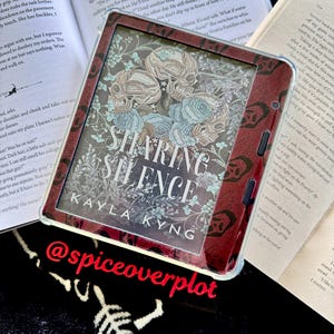 Op de afbeelding: Een vierkante e-reader met een donkerrode en zwarte beschermhoes. Het scherm toont de boekomslag van Sharing Silence door Kayla Kyng, met een illustratie van schedels en blauwe rozen. Het apparaat is voorzien van knoppen aan de zijkant.