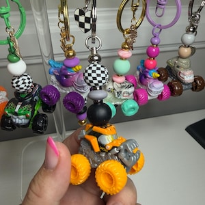May include: A collection of monster truck keychains featuring miniature toy trucks attached to metal rings. Each keychain includes colorful beads, including checkered patterns, and small charms like hearts or lightning bolts. The trucks have oversized wheels and detailed bodies in various colors like orange, green, and purple.