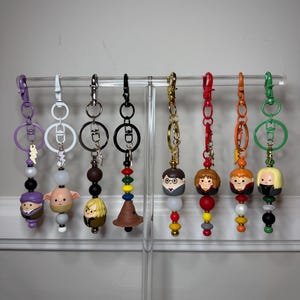 May include: Eight character-themed keychains hanging on a clear stand. Each features a unique figurine head, colorful beads, and a metal clasp. The designs include various figures with distinct hair colors, outfits, and accessories like lightning bolts.
