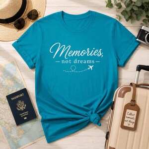 May include: A bright teal short-sleeved t-shirt featuring the white text "Memories, not dreams" with a dashed flight path line and a small aeroplane icon. The shirt has a crew neckline and is made of a solid-coloured fabric with a simple, clean design.