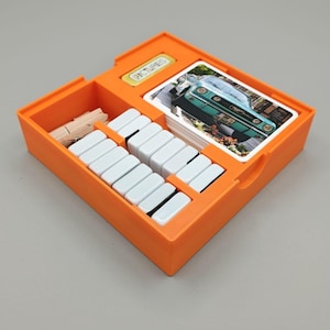 Könnte beinhalten: Ein orangefarbener Brettspiel-Organizer aus Kunststoff mit einem Kartenstapel, der ein türkisfarbenes Auto zeigt, einem kleinen gelben Plättchen mit dem Wort PICTURES, weißen rechteckigen Spielsteinen und hölzernen Wäscheklammern in Fächern.