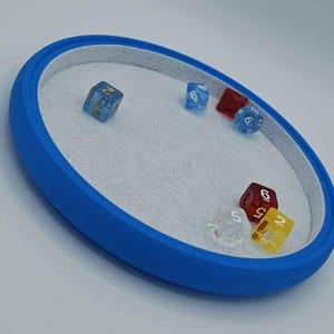 Könnte beinhalten: Ein ovales blaues Würfeltablett mit einer hellgrauen Filzeinlage. Sechs mehrseitige Spielwürfel in verschiedenen Farben, darunter Blau, Rot, Gelb und Transparent, sind auf der Oberfläche des Tabletts verteilt und zeigen jeweils weiße Zahlen.