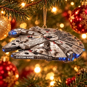 Puede incluir: Un adorno de nave espacial Millennium Falcon en tono plateado con detalles desgastados, una antena parabólica circular y luces LED azules. El lateral del casco incluye una placa rectangular con el texto CUSTOM NAME en negro.