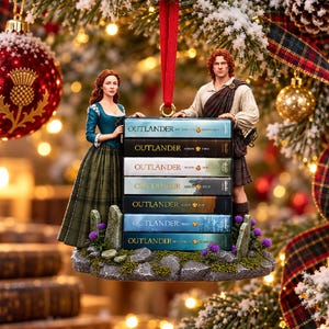 Puede incluir: Un adorno navideño colgante con dos figuras sobre una base rocosa con flores moradas. Las figuras flanquean una pila de siete libros etiquetados como Outlander, con las temporadas de la uno a la siete impresas en letras doradas.