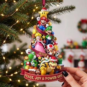May include: A Christmas tree-shaped ornament featuring Mario, Luigi, Princess Peach, Yoshi, Toad, and Bowser. The design includes gold coins, question blocks, and a yellow star at the top. A red ribbon displays the name JESSIE and PEACE, JOY & POWER-UPS.