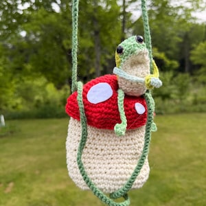 May include: A crochet mushroom-shaped pouch with a cream-colored base and a red cap featuring white felt spots. A small, light green crocheted frog is attached to the top of the mushroom, and the pouch includes a long, thin green crochet strap.