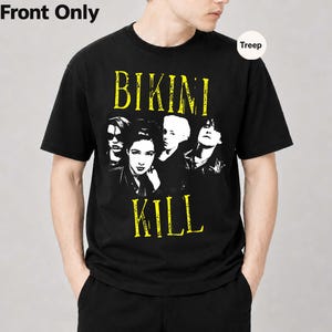 Puede incluir: Una camiseta negra de manga corta con un estampado gráfico en blanco y negro de cuatro personas. El texto BIKINI está impreso en amarillo sobre las figuras, y el texto KILL está impreso en amarillo debajo de las figuras con una fuente desgastada.