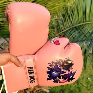 Puede incluir: Un par de guantes de boxeo de color rosa claro con el texto HEN DOG en la muñequera. Los guantes están decorados con un estampado gráfico de tres criaturas moradas de dibujos animados, una con una lengua larga y otra con una sonrisa.