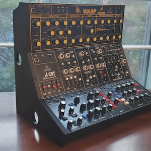 May include: A black desktop synthesiser featuring a top panel labelled WASP DELUXE with yellow knobs and a lower panel labelled CAT with black, blue, and red knobs, sliders, and switches. The unit has a slanted control interface with multiple inputs.