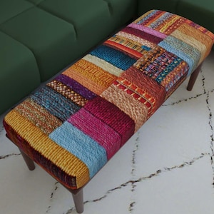 Pode incluir: Um banco retangular com estofamento de retalhos multicolorido e aparência tecida texturizada. O tecido exibe vários blocos com padrões em amarelo, azul, vermelho, laranja e roxo. Apoia-se sobre quatro pernas de madeira castanho-escuras.