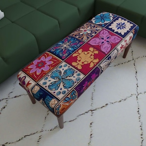 Könnte beinhalten: Eine rechteckige, gepolsterte Bank mit einem lebendigen Patchwork-Muster aus quadratischen Kacheln in Blau-, Rot-, Lila- und Goldtönen. Die Bank steht auf vier dunkelbraunen Holzbeinen und zeigt eine Vielzahl von floralen und geometrischen Motiven.