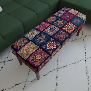 May include: A rectangular ottoman featuring a vibrant, multi-colored patchwork pattern of floral and geometric motifs in shades of blue, red, yellow, and purple. The piece is supported by four dark brown wooden legs and has a cushioned top surface.