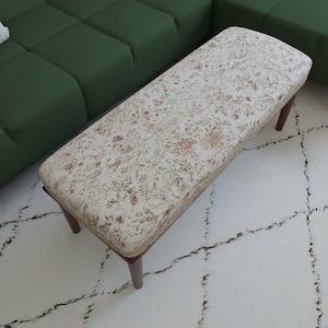 May include: A rectangular ottoman featuring a cream-coloured fabric upholstery with a detailed floral pattern in shades of pink, green, and brown. The piece is supported by four dark brown wooden legs, providing a stable base for the cushioned top.