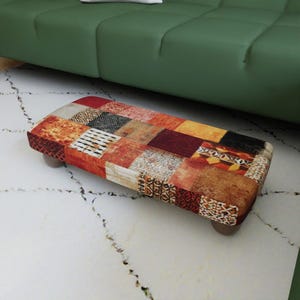 May include: A rectangular footstool featuring a patchwork design with various fabric squares in shades of red, orange, yellow, brown, and black. The upholstery displays a mix of solid colors and intricate geometric patterns, supported by four low, dark feet.