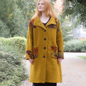 Puede incluir: Un abrigo de color amarillo mostaza con mangas largas, cuello alto, cierre de botones frontal y dos grandes bolsillos de parche con un patrón floral oscuro. El abrigo se lleva sobre un vestido negro y medias oscuras, junto con botines marrones.