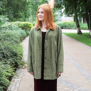 May include: A long-sleeved olive green textured jacket with a collar and front buttons worn over a floor-length dark burgundy dress and black ankle boots. The jacket features a subtle speckled pattern and a straight hemline that reaches the hips.