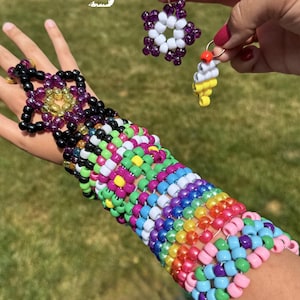 Può includere: Un grande bracciale a fascia composto da perline di plastica multicolori intrecciate in un motivo complesso nei toni del viola, verde, giallo, blu, rosa e arancione, accompagnato da un portachiavi a forma di stella e un piccolo ciondolo.