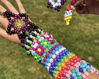 Può includere: Un grande bracciale a fascia composto da perline di plastica multicolori intrecciate in un motivo complesso nei toni del viola, verde, giallo, blu, rosa e arancione, accompagnato da un portachiavi a forma di stella e un piccolo ciondolo.