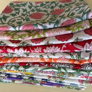 May include: A stack of folded rectangular fabric napkins featuring various floral patterns in vibrant colors including pink, red, orange, yellow, green, and purple on a white background. Each piece of cloth displays distinct botanical prints.