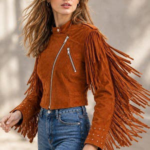 May include: A rust-colored suede jacket featuring long fringe detailing along the sleeves and back. The jacket has a stand-up collar with a snap closure, an asymmetrical silver-tone zipper, zippered chest pockets, and silver stud accents on the cuffs.