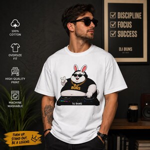 Peut inclure: Un t-shirt blanc coupe oversize avec une impression graphique représentant un lapin de dessin animé portant des lunettes de soleil, un casque et une chaîne en or, mixant sur une console. Le texte DJ BUNS apparaît sur le t-shirt et la console.