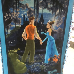 May include: A blue fleece blanket with a printed image of a man and woman standing in front of a castle. The image is set against a dark blue background with trees and a moon. The text "Disney Princess" is printed in the bottom right corner.
