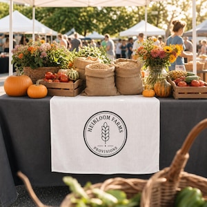 Puede incluir: Un camino de mesa blanco con un logotipo circular negro que dice Heirloom Farms Provisions y un icono de espiga de trigo. La mesa muestra dos sacos de arpillera con granos, cajas de madera con tomates rojos, calabazas naranjas y jarrones con flores.