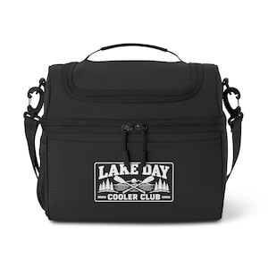 Puede incluir: Una bolsa térmica negra con asa superior y correa ajustable para el hombro. El frente presenta un estampado gráfico blanco con el texto LAKE DAY COOLER CLUB, que muestra remos cruzados, pinos y un sol sobre un cuerpo de agua.