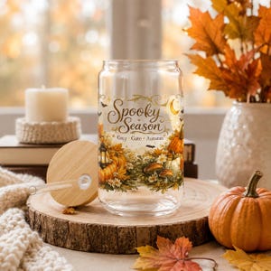 May include: A clear glass tumbler with a bamboo lid and glass straw, featuring a printed design of pumpkins, autumn flowers, a lantern, bats, and the text Spooky Season Cozy Cute Autumn in gold and orange tones on the exterior surface.