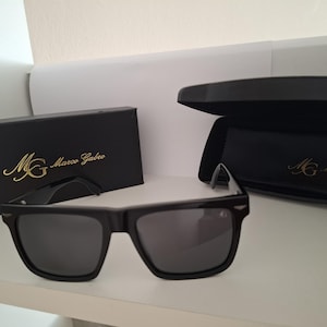 Könnte beinhalten: Schwarze Sonnenbrille mit dunklen Gläsern und rechteckigem Rahmen. Das Set enthält eine schwarze rechteckige Schachtel und ein schwarzes Hartschalenetui, beide mit dem goldfarbenen Logo und dem Schriftzug Marco Gabro auf der Vorderseite.