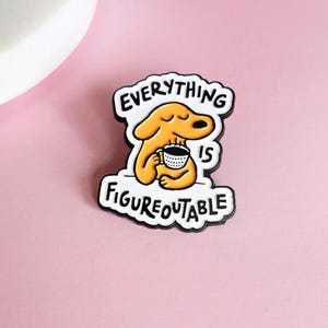 May include: A metal enamel pin featuring a cartoon orange dog holding a white coffee mug with black polka dots. The pin has a white background with black outlines and the black text "EVERYTHING IS FIGUREOUTABLE" written around the dog.