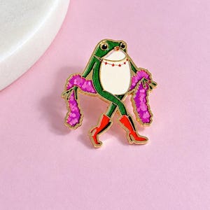 May include: A gold-toned enamel pin featuring a green frog with a white belly, wearing red boots and a red star-patterned necklace. The frog is draped in a bright pink feather boa, with its legs crossed in a pose, outlined in gold metal.