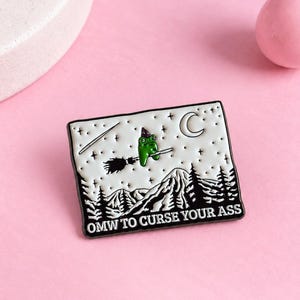以下が含まれることがあります： 白い背景に黒い山脈と森のシルエットが描かれた長方形のエナメルピン。紫色の帽子をかぶった緑色のカエルが、三日月と星の下でほうきに乗って飛んでいる様子がデザインされています。テキストは「OMW TO CURSE YOUR ASS」と書かれています。