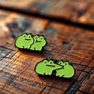 May include: Two lime green enamel pins featuring cartoon frogs. One pin shows two frogs sitting side by side, while the other pin depicts one frog with a long, pointed snout touching the face of a second frog. Both pins have black metal outlines.