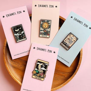Può includere: Quattro spille in smalto rettangolari su cartoncini di supporto individuali. Le spille presentano illustrazioni in stile tarocchi intitolate The Reader, The Sun, The Coffee e The Moon, con scheletri, corpi celesti e motivi floreali dettagliati.