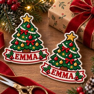 May include: Two embroidered Christmas tree patches featuring green foliage, red and gold ornaments, white garland, and a yellow star topper. Each patch has a white banner at the base with the name EMMA in red letters and holly accents.