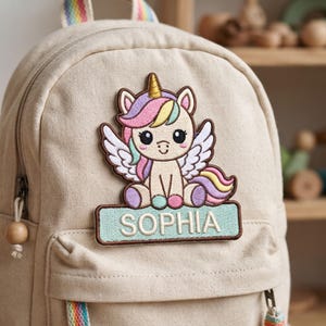 May include: A beige canvas backpack featuring an embroidered patch of a winged unicorn with a multicolored mane and tail. Below the unicorn, a light blue rectangular patch displays the name SOPHIA in white capital letters with a brown border.