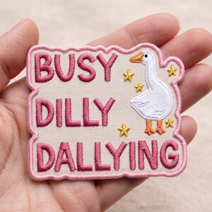 May include: A rectangular embroidered patch with a light beige background and a pink border. It features the dark pink text BUSY DILLY DALLYING, a white goose with an orange beak and feet, and several small yellow embroidered stars.