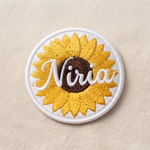 May include: A circular embroidered patch featuring a yellow sunflower with a dark brown center. The name Niria is stitched across the center in white cursive script. The patch has a white border and a dense, textured thread finish throughout.