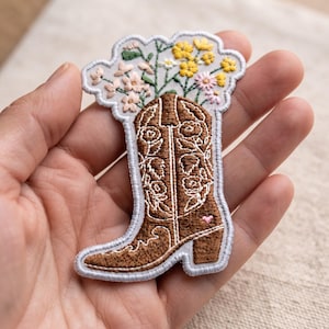 May include: An embroidered patch shaped like a brown cowboy boot with white floral stitching details. The boot is filled with a bouquet of embroidered pink, white, and yellow wildflowers. The patch has a light gray border and a small pink heart.