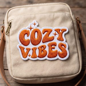 May include: A beige canvas crossbody bag featuring an embroidered patch on the front. The patch displays the text COZY VIBES in bold, orange, rounded letters with small star accents, all outlined in white thread against a white background.
