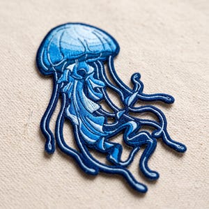 May include: An embroidered patch in the shape of a jellyfish, featuring a gradient of blue and white threads. The design displays a rounded bell top with intricate stitching and multiple long, flowing tentacles extending downward in dark blue.