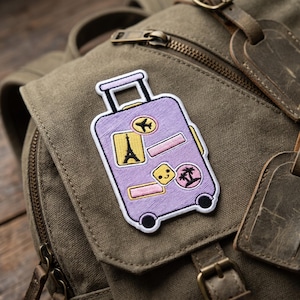 May include: An embroidered patch shaped like a light purple suitcase with a retractable handle and two black wheels. The suitcase features several small travel-themed stickers, including an airplane, the Eiffel Tower, palm trees, and a smiling face.