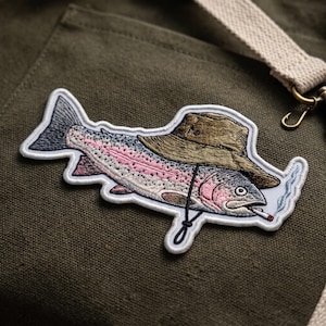 May include: An embroidered patch featuring a rainbow trout wearing a brown bucket hat with a chin strap. The fish has a pink lateral stripe, grey scales, and is holding a lit cigarette in its mouth with a small trail of blue smoke rising.