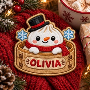 May include: An embroidered patch featuring a white dumpling character with a carrot nose, black top hat, and red scarf, sitting inside a tan bamboo steamer basket. The text OLIVIA is stitched in red on the front, flanked by two blue snowflakes.