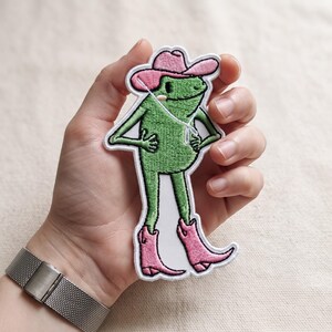 May include: An embroidered patch featuring a green frog character wearing a pink cowboy hat and matching pink cowboy boots. The frog has a white string detail hanging from the hat and is depicted in a standing pose with hands on its hips.