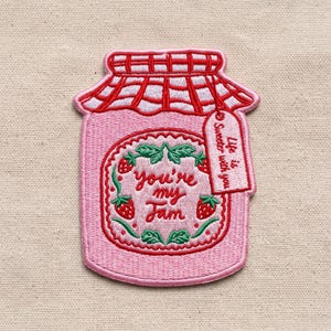 May include: An embroidered patch shaped like a pink jam jar with a red checkered lid. The center features red strawberries and green leaves with the text "you're my Jam". A small tag attached to the lid reads "Life is sweeter with you".