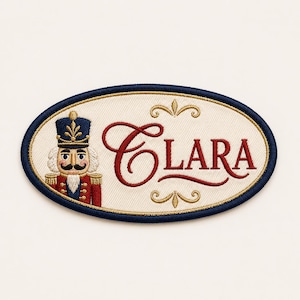 May include: An oval embroidered patch with a cream-colored background and a dark blue border. The patch features a nutcracker figure on the left and the name Clara embroidered in red script, accented by gold decorative flourishes above and below.