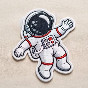 May include: An embroidered iron-on patch featuring a white astronaut suit with red accents, a black helmet visor with white highlights, and a chest control panel with red and grey buttons. The astronaut is depicted in a floating pose with one arm raised.